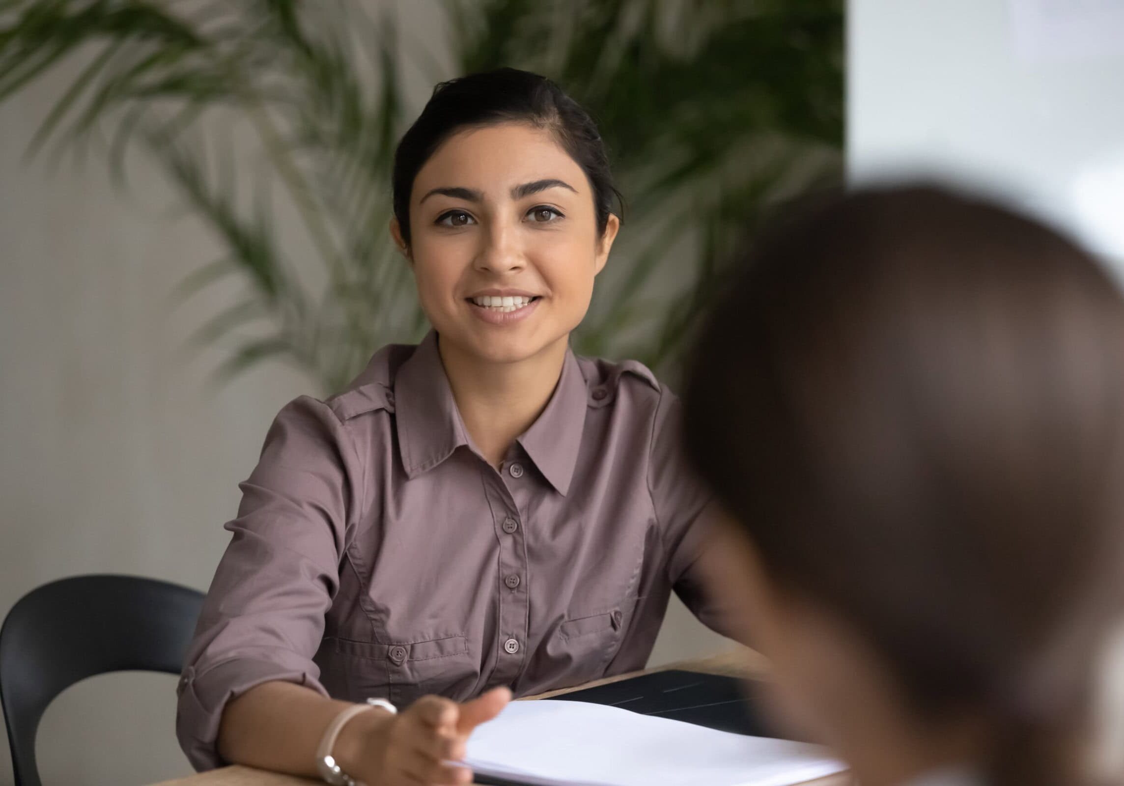 icf-become-a-coach-female-coach-educator Smiling woman in a violet blouse sitting at a desk and reviewing coaching materials with a student in a modern office.