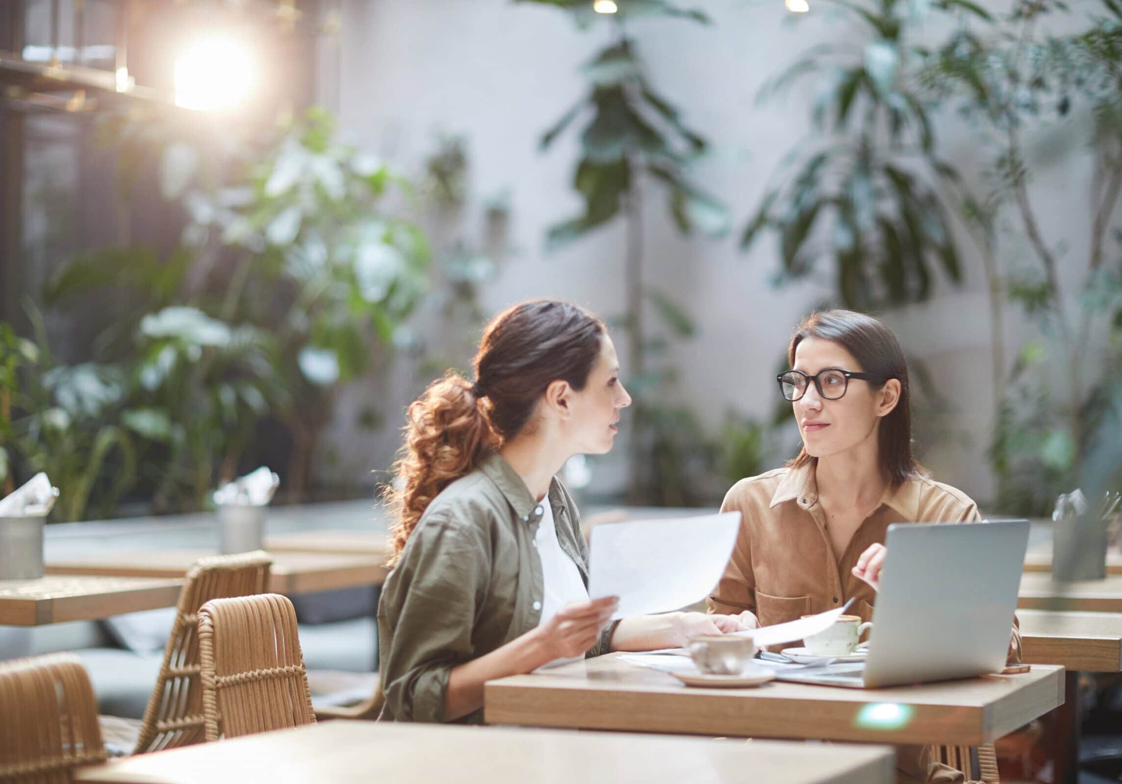 Two Young Businesswomen in Cafe An experienced coach in glasses is meeting with an aspiring coach in an empty sunlit cafe with plants, reviewing coaching materials, and using a laptop.