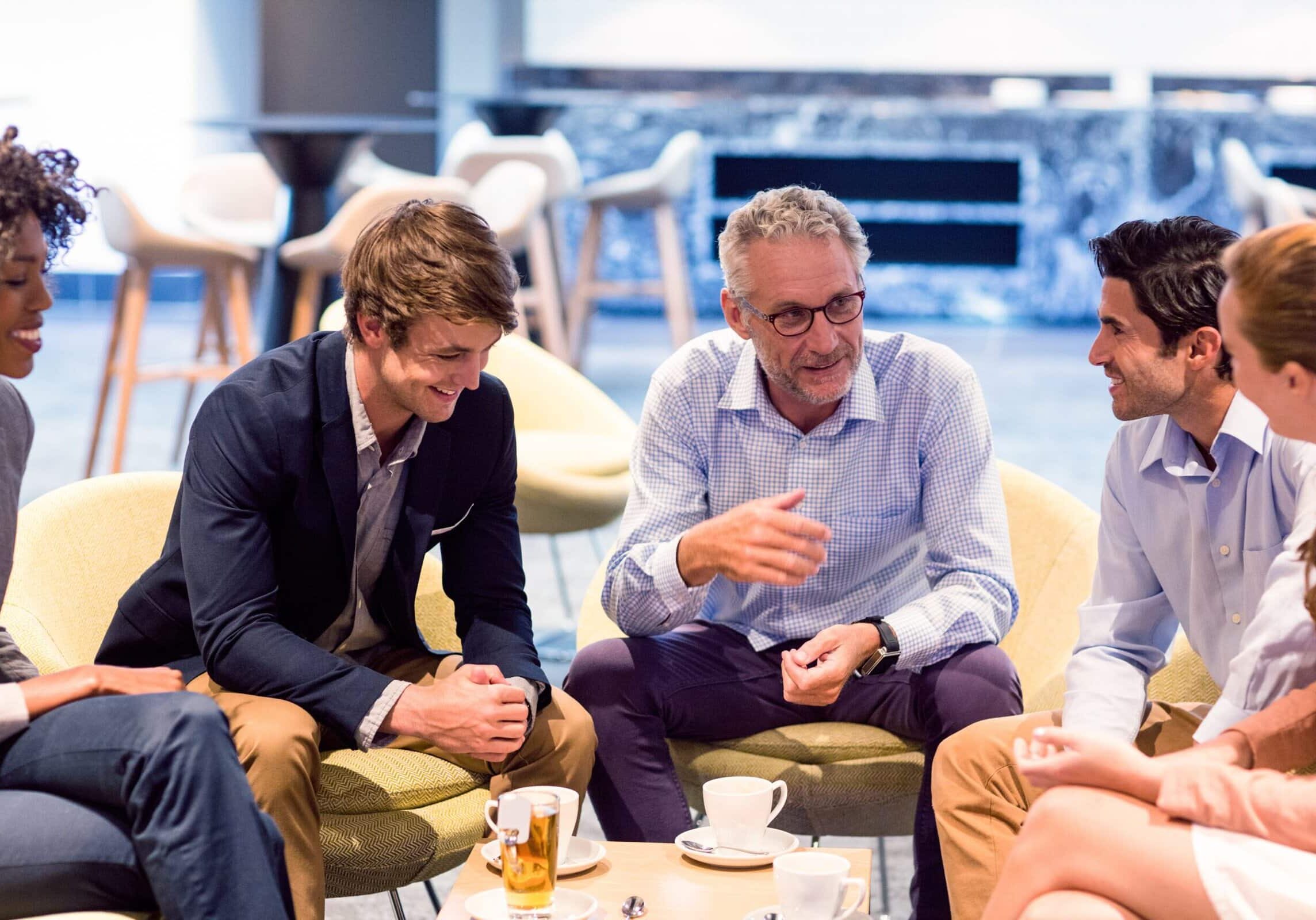 Coaching Group Circle A group of five professionals seated in a lounge, discussing coaching and smiling over coffee, fostering teamwork and collaboration.