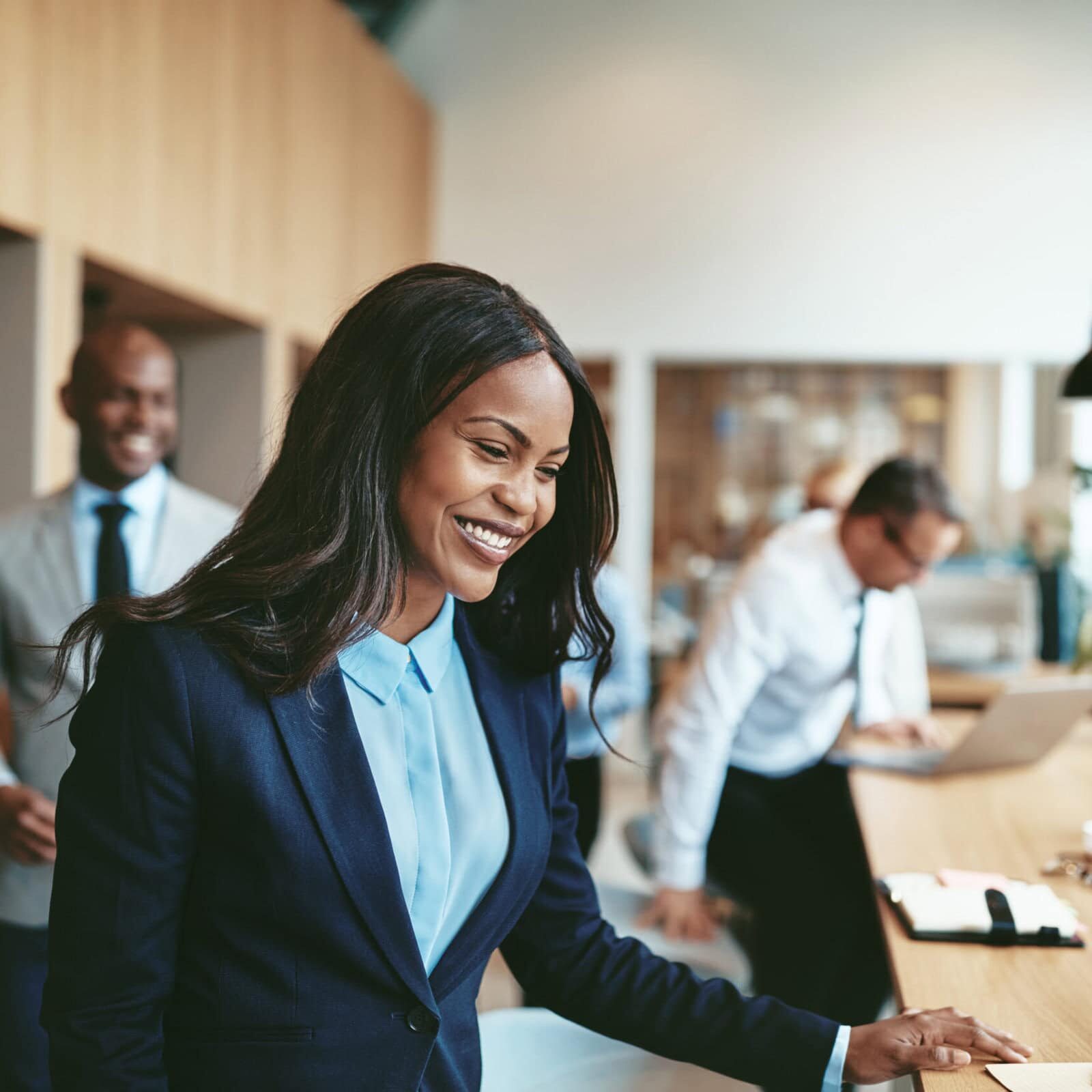 Smiling African American businesswoman sitting down for an office meeting A woman is in the focal point, standing behind a desk smiling, while other co-workers are moving to sit at the same desk.